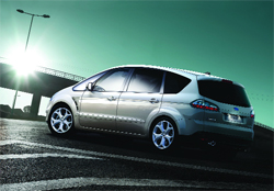 A silver minivan parked on dark asphalt with a bright sun and bridge in the background.