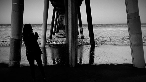 Silhouette of a person standing on a beach under the wooden pilings of a long pier at dusk.