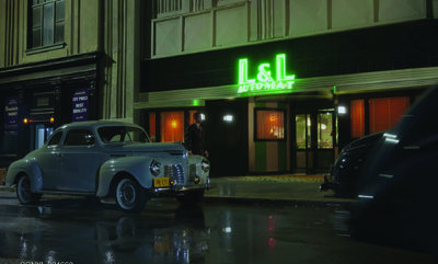 A light blue vintage car parked on a wet street in front of a glowing L&L Automat storefront at night.