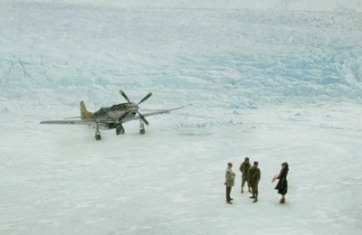 A vintage propeller plane rests on a vast, white ice field near four people standing in the distance.