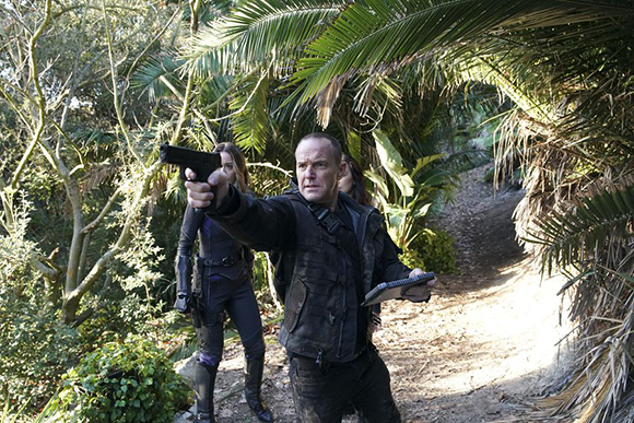 A man in a dark jacket aims a handgun while walking on a forest path with a woman behind him.