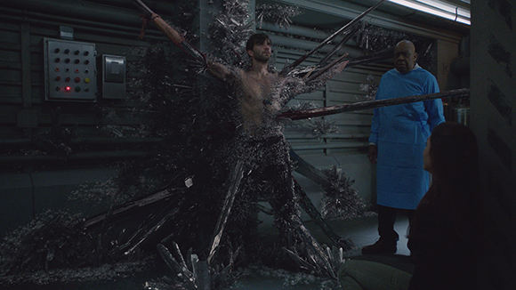 A man is pinned to a wall by long, sharp metallic shards while a man in blue scrubs watches.