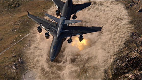 A dark military cargo plane flies low over a rocky terrain, kicking up a massive cloud of dust and debris.