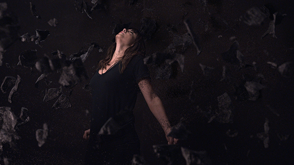 A woman in dark clothing lies amidst floating dark fragments in a dark space.