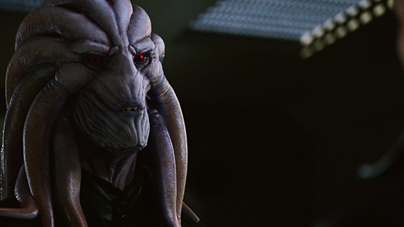 Close-up of a grey-skinned alien with large red eyes and thick, tentacle-like facial appendages.