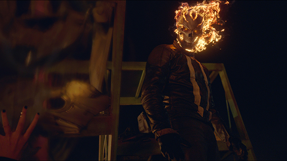 A figure in a dark leather jacket with a skull head engulfed in bright orange flames stands against a dark background.
