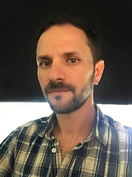 A man with short dark hair and a beard wearing a plaid shirt against a dark background.