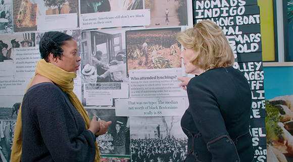 A woman in a yellow scarf speaks to a blonde woman in front of a wall of historical displays.