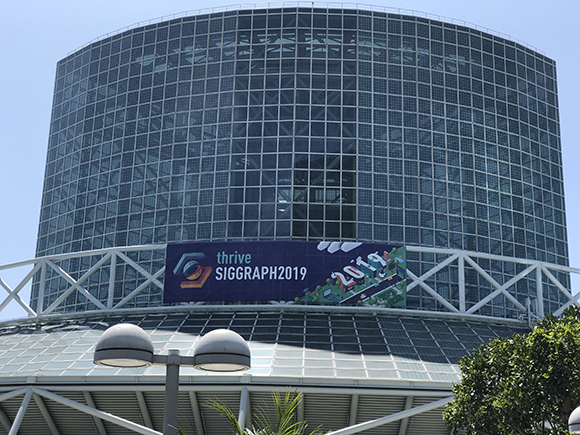 A banner reading "thrive SIGGRAPH 2019" is hung on a large, curved glass building with a white lattice structure.