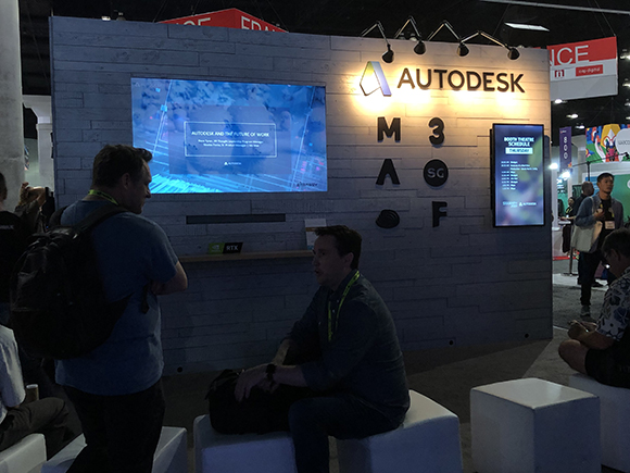 Two men are at an Autodesk booth at a trade show. One man stands with a backpack, and another man sits on a white cube.