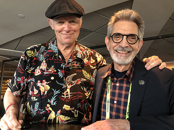 Two men smile at the camera. The man on the left wears a black Hawaiian shirt with colorful graphics and a dark cap. The man
