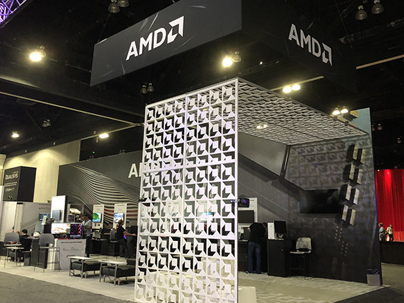 AMD logo displayed prominently on hanging banners and booth graphics at a trade show. Interactive computer displays are set u