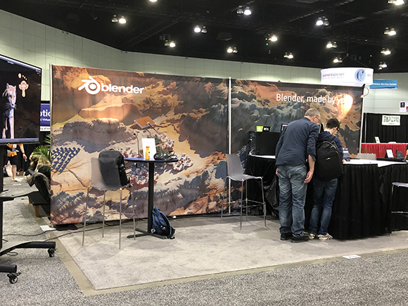 Two people stand at a Blender booth, looking at a computer screen. The booth has a large backdrop with the Blender logo and a