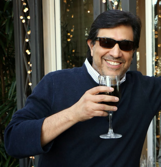 A smiling man in sunglasses and a navy sweater holds a wine glass filled with a dark beverage.