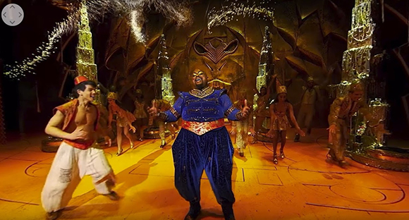 A performer in a blue ornate costume stands center stage among dancers in a theatrical production.