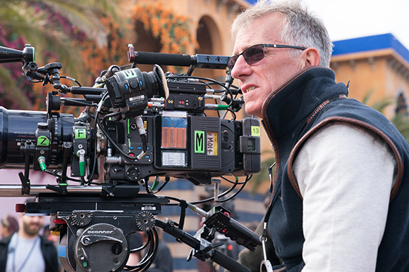 A man with grey hair and sunglasses looks through the viewfinder of a large professional film camera.
