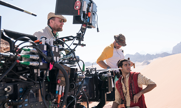A film crew operates a large camera rig in a sunny desert with sand dunes and mountains.