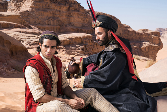 A man in a red vest sits while another man in a black robe and turban leans toward him in a desert.