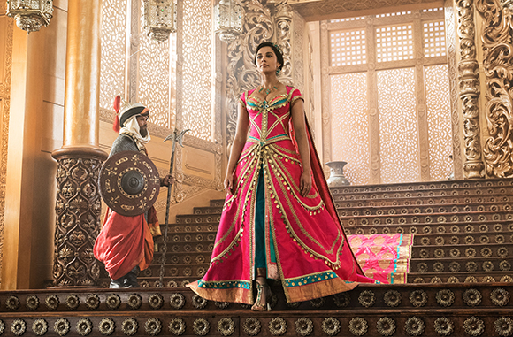 A woman in an ornate pink gown stands on a grand staircase beside a man holding a shield and staff.