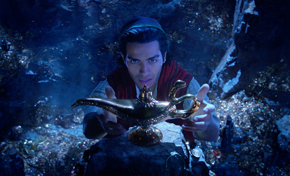 A man in a red vest reaches toward a golden oil lamp resting on a rock amidst a cave of jewels.