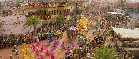 A wide aerial view of a crowded street parade with large floats, elephants, and people in colorful costumes.