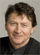 Close-up headshot of a middle-aged man with wavy brown hair, wearing a dark suit and tie, smiling.