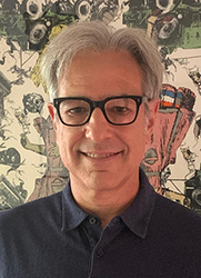 A man with gray hair and black-rimmed glasses smiling in front of a patterned background.
