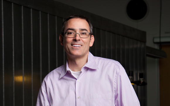 A man with glasses and a light purple striped shirt smiling in a studio setting.
