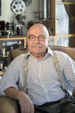 Elderly man with glasses and a mustache wearing a striped shirt and suspenders, sitting in an armchair.