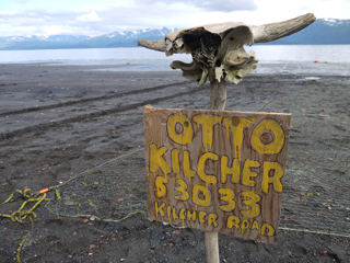 A wooden sign reading 'OTTO KILCHER 53033 KILCHER ROAD' with an animal skull mounted on a post on a dark beach.