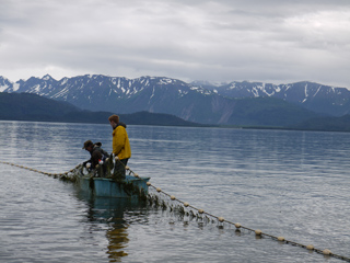 Two people in a small blue boat pull in a fishing net on a calm lake with snow-capped mountains behind them.