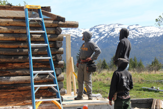 Three people in hooded clothing stand near a log cabin under a snowy mountain range.