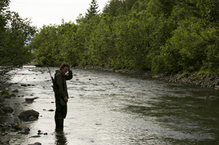 A person in dark clothing stands in a shallow river holding a rifle, surrounded by dense green trees.