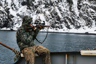 A person in camouflage gear sits on a boat, aiming a scoped rifle toward snowy cliffs across the water.