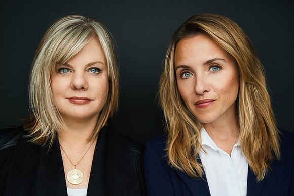 Two women with blonde hair looking at the camera against a dark backdrop.