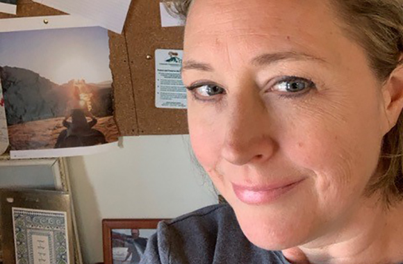 Close-up of a woman smiling slightly in front of a corkboard with various photographs.