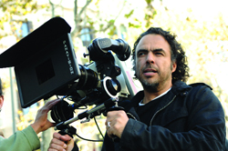 A man with dark curly hair looks intently while holding the handles of a large Panavision film camera.