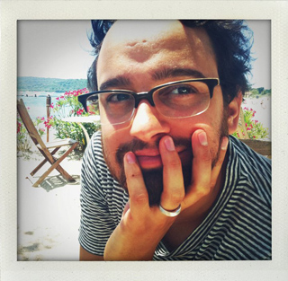 A man with dark hair, glasses, and a beard rests his hand on his face in a sunny outdoor setting.