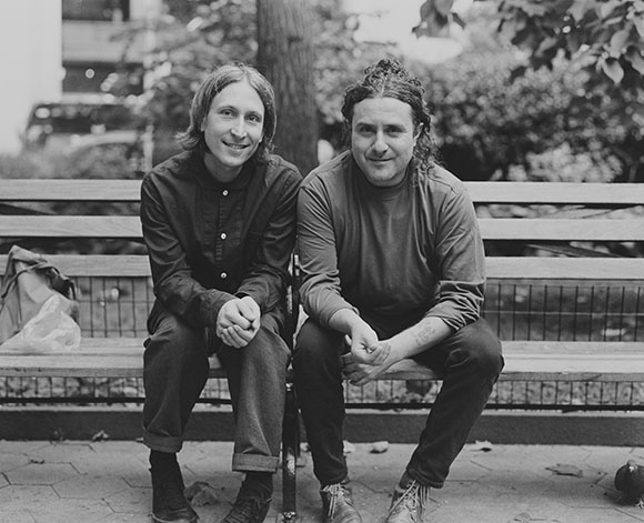 Two men sit side-by-side on a wooden park bench outdoors.