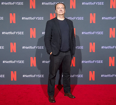 Man in a dark blazer and navy shirt posing on a red carpet against a Netflix branded wall.