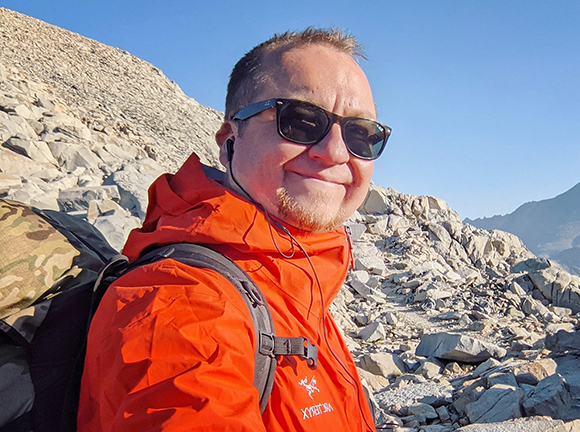 A man wearing sunglasses and an orange jacket smiles in a rocky, mountainous area.