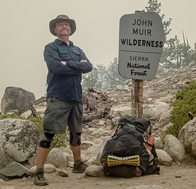 A man in hiking gear stands with arms crossed next to a John Muir Wilderness sign and a backpack.