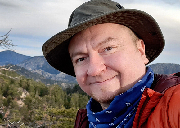 A man wearing a brown hat and blue bandana smiling in a mountainous landscape.
