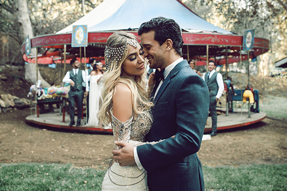 A man in a dark suit and a woman in a beaded gown embrace in front of a red carousel.