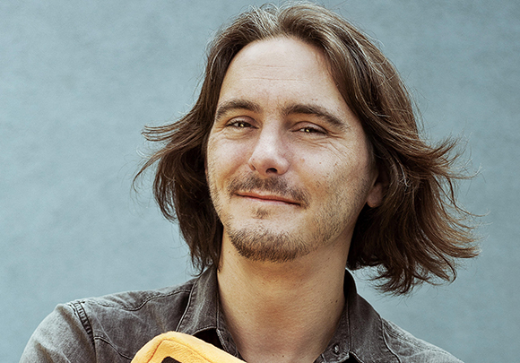 A man with medium-length brown hair and a light beard smiling against a gray background.