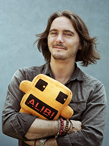 A man with shoulder-length hair smiling while holding an orange plush toy that says ALIBI.