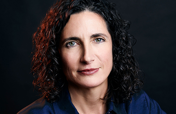 A woman with dark curly hair and green eyes looks forward against a dark background.