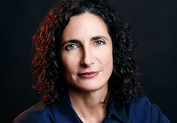 A close-up portrait of a woman with dark curly hair and light eyes looking directly at the camera.