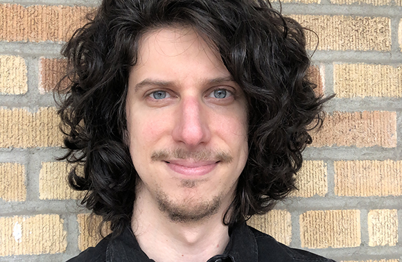 A person with long, dark curly hair and blue eyes smiling against a tan brick wall.