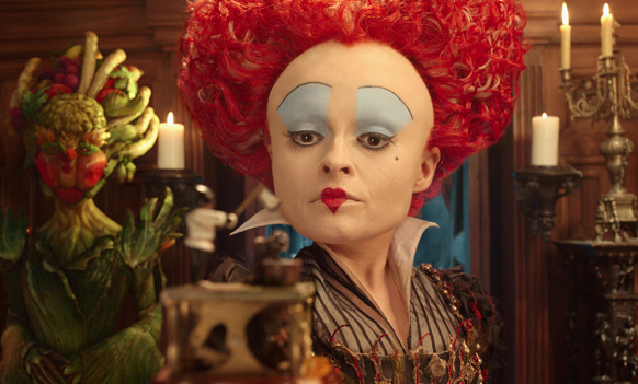 A woman with large red hair, blue eyeshadow, and red lipstick looks forward in a dimly lit room.